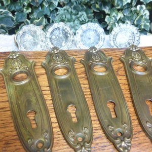 May include: Five antique brass door backplates with ornate designs and keyholes. The backplates are arranged in a row on a wooden surface.
