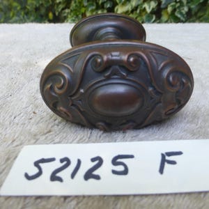Può includere: Un pomello per porta antico in bronzo con forma ovale e un ornato design a volute. Il pomello ha una finitura marrone scuro ed è probabilmente in metallo. Il numero "52125 F" è scritto su un'etichetta bianca sottostante.