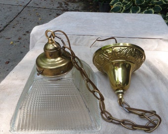 Op de afbeelding: Twee goudkleurige lampen met heldere glazen lampenkappen. De ene kap is vierkant en de andere rond. Beide kappen hebben een geribbelde textuur. De lampen zijn met een gouden ketting verbonden.