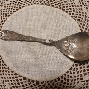 Puede incluir: Una cuchara plateada con detalles ornamentales de uvas y hojas en el cuenco y el mango. La cuchara descansa sobre un paño blanco y circular, sobre un fondo de un tapete crema de tejido abierto.