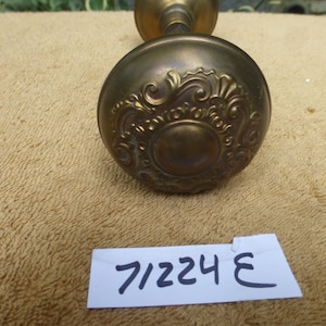 Puede incluir: Una perilla de puerta de latón con un diseño ornamentado. La perilla es redonda y tiene un centro elevado. La perilla está unida a un eje de latón. El texto "7/224 E" es visible en una etiqueta blanca.