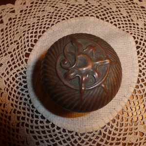 May include: A round, antique bronze-colored container with a sunburst design on the lid. The container sits on a white crocheted doily, with a detailed lace pattern. The container's lid has a spiral and leaf design.