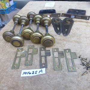 May include: A collection of vintage brass door hardware, including several doorknobs, strike plates, and a latch. The doorknobs have a classic round design. Also included are screws and other related components. The label reads "101625 A".