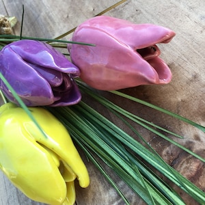 以下が含まれることがあります： 紫、ピンク、黄色のセラミック製のチューリップ3本の花束。チューリップは、緑色の茎と葉のある束にまとめられています。