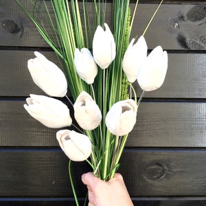 A bouquet of 9 handmade white ceramic tulips with green stems and leaves. The tulips are arranged in a hand-held bouquet against a dark black wooden panel wall.