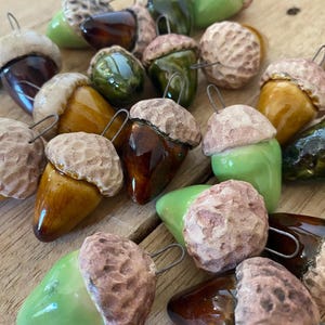 May include: A collection of ceramic acorn ornaments featuring glossy, smooth, teardrop-shaped bodies in shades of light green, dark green, and brown. Each acorn has a textured, matte-finish cap in a light tan colour and a metal wire loop for hanging.