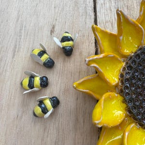 May include: A close-up of a ceramic sunflower with yellow petals and a dark center, alongside four small, detailed bee figurines. The bees are yellow and black with white wings, set against a wooden background.