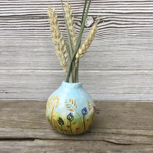 May include: A small, round, hand-painted ceramic vase with a light blue background and a wheat field design. The vase holds dried wheat stalks and green stems. The vase is sitting on a wooden surface.