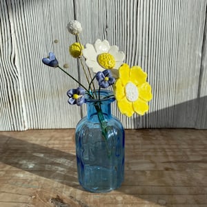 May include: A small blue glass bottle containing a bouquet of handcrafted ceramic flowers. The arrangement features two large blossoms in yellow and white, alongside smaller purple buds and a textured white sphere on thin green wire stems.