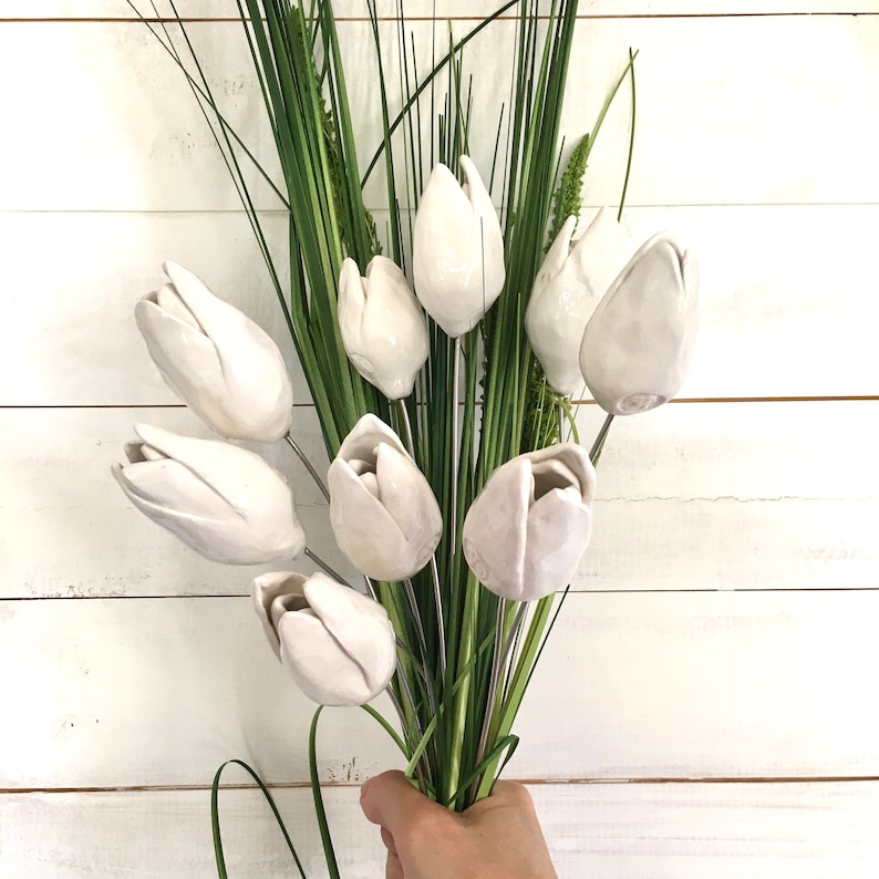 A bouquet of 9 handmade white ceramic tulips with green stems and leaves. The tulips are arranged in a hand-held bouquet against a white wooden panel wall.