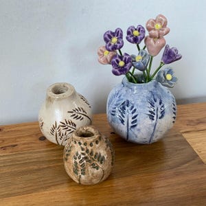 Könnte beinhalten: Drei kleine Keramikvasen mit Blumendesigns. Die Vase in der Mitte ist blau und weiß und enthält einen Strauß aus lila, rosa und blauen Keramikblumen. Die beiden anderen Vasen sind braun und weiß mit einem Blattdesign.