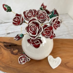 May include: A bouquet of red ceramic roses with white accents in a white ceramic vase. The vase is sitting on a wooden surface with a white ceramic heart that says "LOVE" on it.