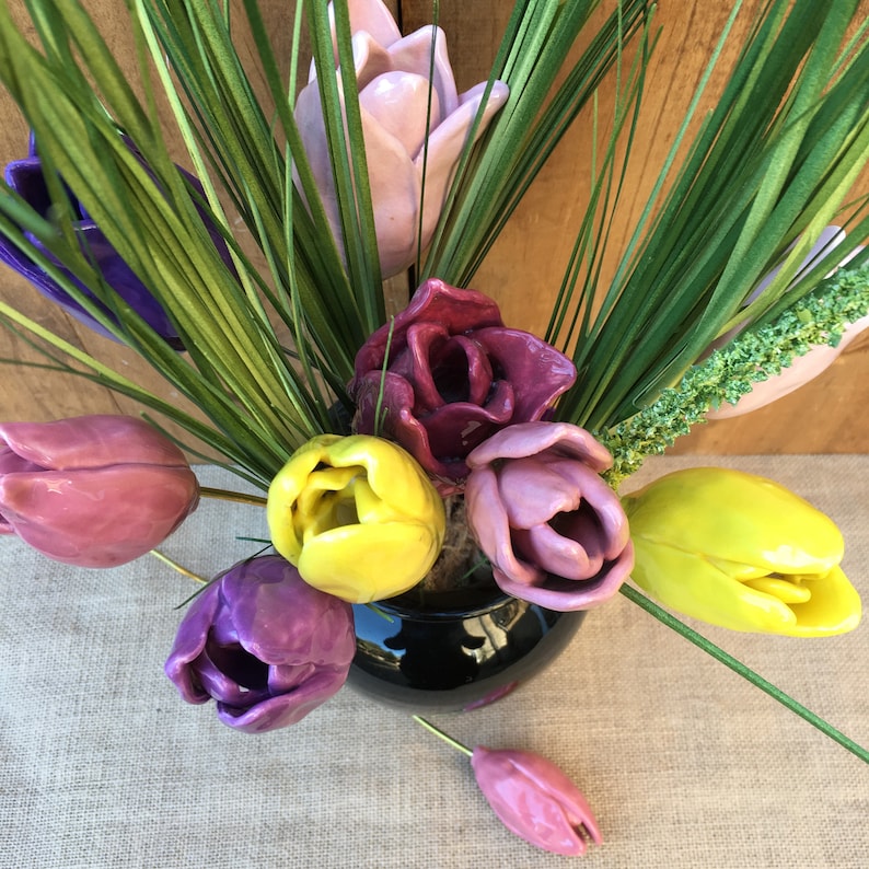 the over view of a bouquet of 9 handmade ceramic tulips in a vase on a table top. The tulips are different bright colours, yellow, pink, lilac, white, purple. the photo taken from above shows the petals of the flowers slightly open in a natural way.