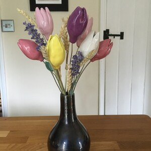 A bouquet of handmade ceramic tulips in a black vase. The tulips are in shades of pink, purple, yellow, and white. The vase is on a wooden table.