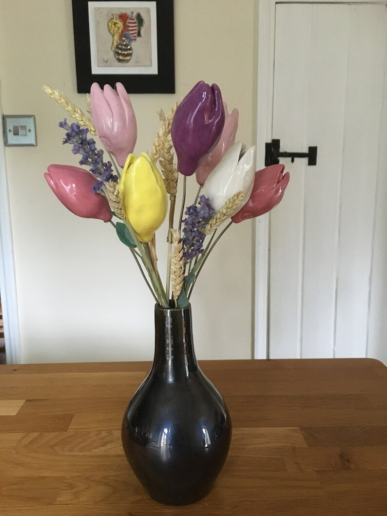 A bouquet of handmade ceramic tulips in a black vase. The tulips are in shades of pink, purple, yellow, and white. The vase is on a wooden table.