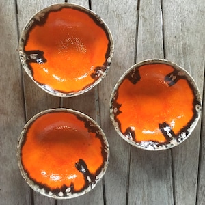 May include: Three ceramic bowls with a bright orange glaze. The bowls have a textured rim with dark brown accents. The interior of each bowl is a glossy orange. The bowls are handmade.