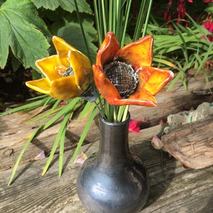 Op de afbeelding: Twee keramische bloemen in een zwarte vaas. De bloemen zijn geel en oranje met een getextureerd midden. De vaas heeft een eenvoudige, ronde vorm.