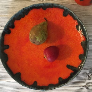 Könnte beinhalten: Ein leuchtend orangefarbener Keramikteller mit einem schwarzen und blauen Rand. Der Teller hat eine strukturierte Oberfläche und zeigt eine Birne und einen Apfel.