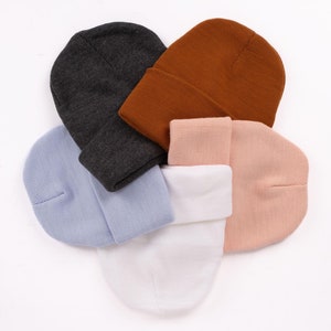 Peut inclure: Quatre bonnets en tricot de différentes couleurs : gris, marron, bleu clair et rose. Les bonnets sont disposés en cercle sur un fond blanc.
