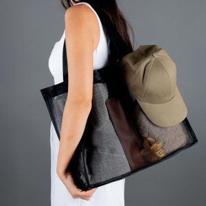 May include: A woman is carrying a black mesh tote bag with a brown leather accent. The bag contains a gray towel, a beige baseball cap, and a wooden decorative item.