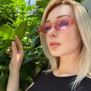 May include: A woman wearing pink rectangular sunglasses with a black t-shirt.