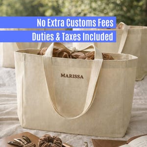 May include: A cream-colored canvas tote bag with two sturdy handles and the name MARISSA embroidered in brown thread on the front. The bag has an open top, revealing brown paper filler inside, and is constructed from a durable woven fabric.