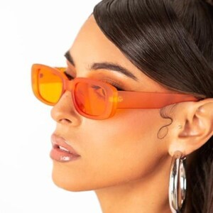 May include: A person wearing orange tinted rectangular sunglasses with a silver hoop earring and a silver chain necklace.