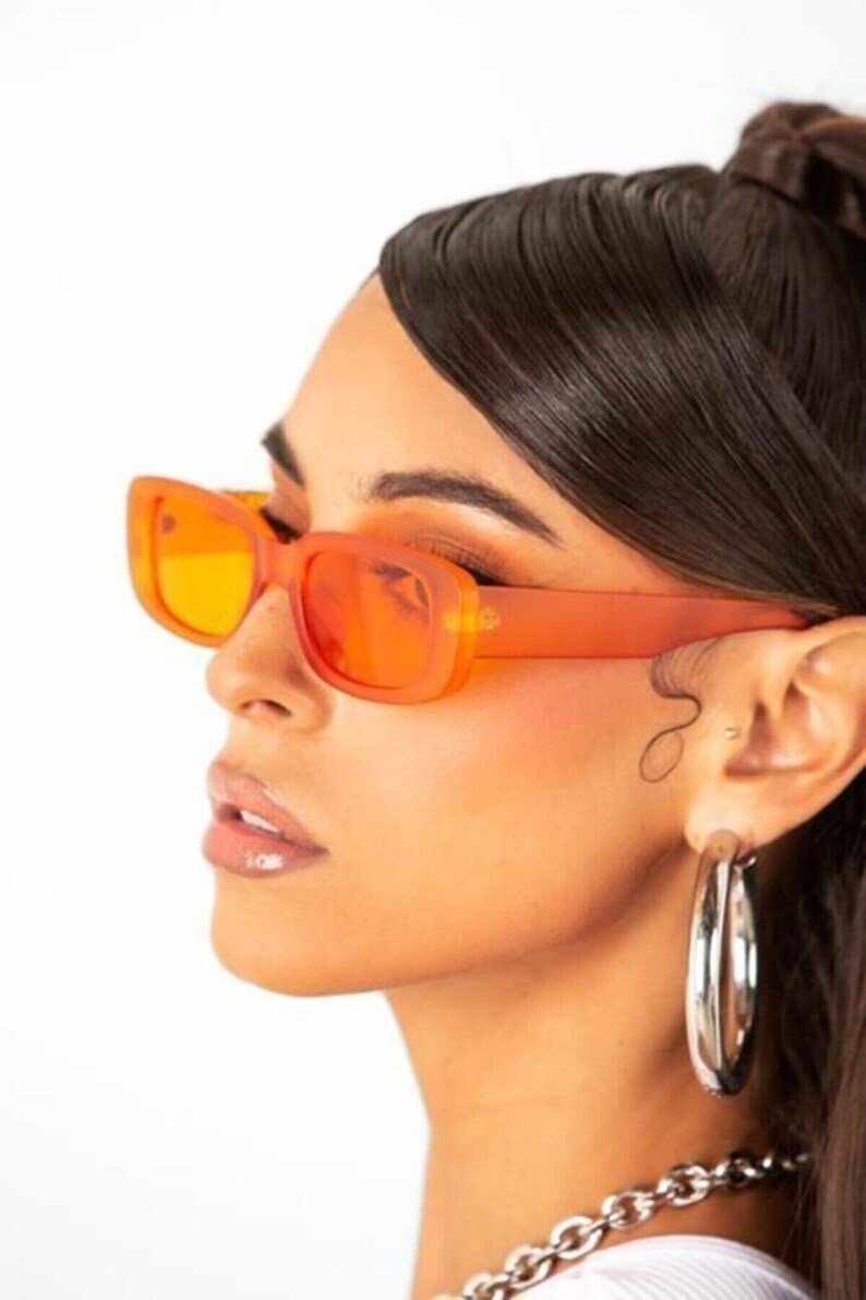 May include: A person wearing orange tinted rectangular sunglasses with a silver hoop earring and a silver chain necklace.