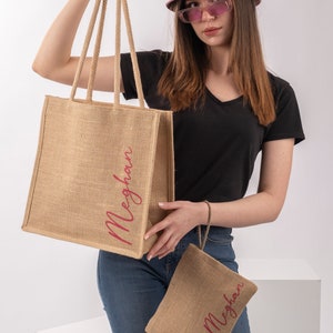 May include: A set of two personalized jute tote bags with the name "Meghan" printed in pink on each bag. The larger bag has two handles and the smaller bag has one handle. The bags are shown in front of a white background.
