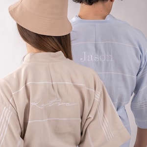 K&ouml;nnte beinhalten: Zwei Personen tragen passende Badem&auml;ntel. Einer ist beige mit wei&szlig;en Streifen und dem Namen "Katie" auf dem R&uuml;cken gestickt. Der andere ist hellblau mit wei&szlig;en Streifen und dem Namen "Jason" auf dem R&uuml;cken gestickt. Ein beiger Fischerhut ist ebenfalls zu sehen.