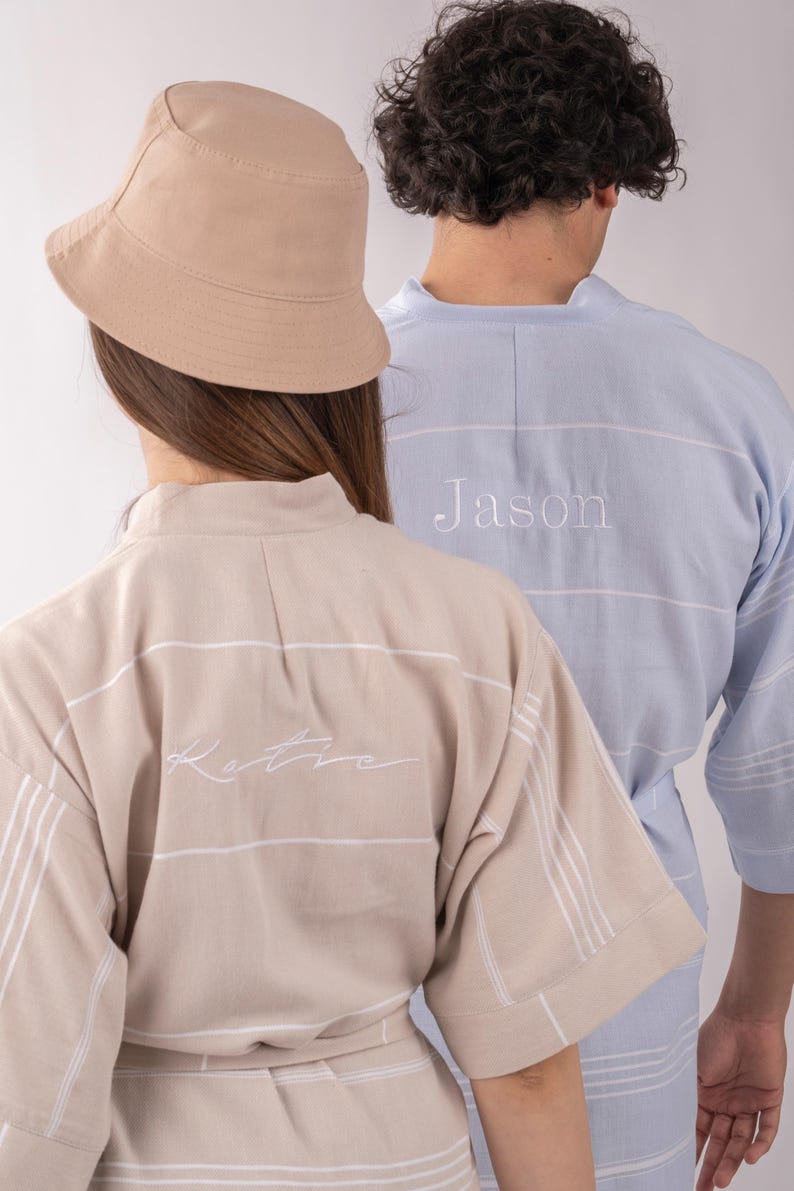 K&ouml;nnte beinhalten: Zwei Personen tragen passende Badem&auml;ntel. Einer ist beige mit wei&szlig;en Streifen und dem Namen "Katie" auf dem R&uuml;cken gestickt. Der andere ist hellblau mit wei&szlig;en Streifen und dem Namen "Jason" auf dem R&uuml;cken gestickt. Ein beiger Fischerhut ist ebenfalls zu sehen.