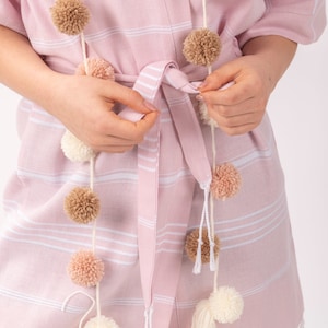 K&ouml;nnte beinhalten: Hellrosa Bademantel mit wei&szlig;en Streifen und Fransen am Saum. Eine Pompon-Girlande in Creme, Beige und Rosa ist &uuml;ber dem Bademantel drapiert. Der Bademantel wird mit einem passenden G&uuml;rtel in der Taille gebunden.