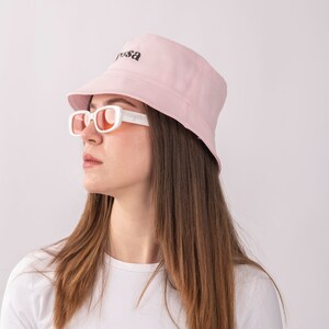 May include: A woman wearing a pink bucket hat with the word "Yrsa" embroidered on it. She is also wearing white sunglasses and a white t-shirt.