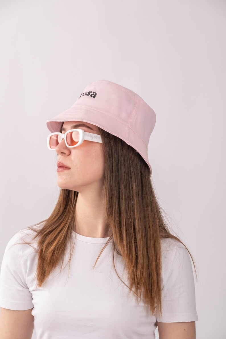 May include: A woman wearing a pink bucket hat with the word "Yrsa" embroidered on it. She is also wearing white sunglasses and a white t-shirt.