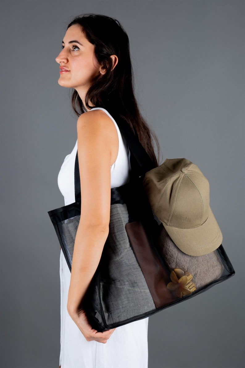 May include: A woman is carrying a black mesh tote bag with a brown leather accent. The bag contains a gray towel, a beige baseball cap, and a wooden decorative item.