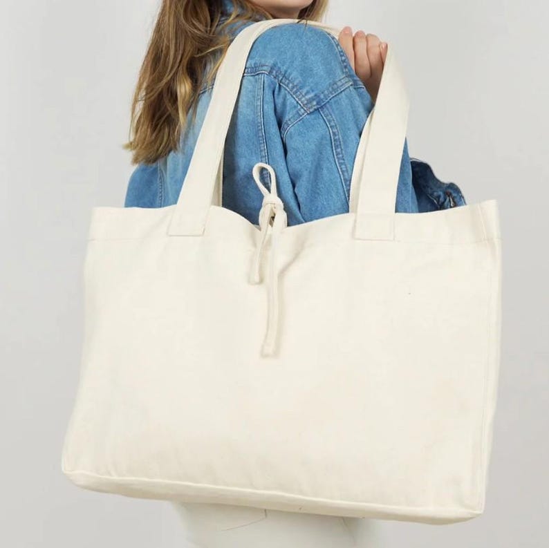 May include: A large, off-white canvas tote bag featuring two wide shoulder straps and a central drawstring closure with a knotted cord detail. The bag has a rectangular shape and a plain, unpatterned surface, constructed from a sturdy fabric material.