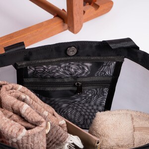 May include: Black mesh tote bag with a zippered pocket and a snap closure. The bag is filled with a brown scarf, a beige towel, and a beige wallet.
