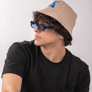 May include: A person wearing a black t-shirt with a pocket and a beige bucket hat with a blue "K" logo. The person is also wearing blue tinted sunglasses with black frames that say "Kevin" on the side.