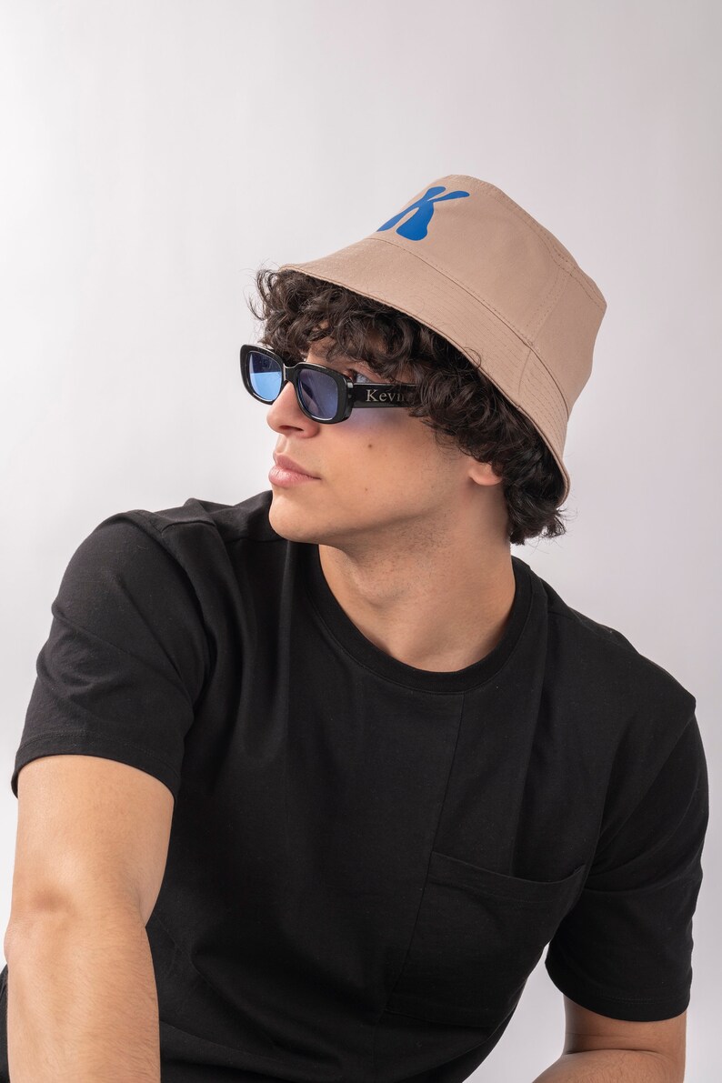 May include: A person wearing a black t-shirt with a pocket and a beige bucket hat with a blue "K" logo. The person is also wearing blue tinted sunglasses with black frames that say "Kevin" on the side.