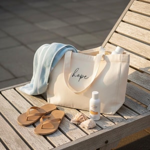 May include: A cream-colored canvas tote bag with the word hope printed in black script, a light blue and white striped towel, a pair of tan flip-flops, a white bottle of sunscreen, and two small seashells arranged on a wooden surface.