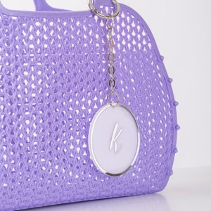 Pu&ograve; includere: Una borsa tote in plastica color lavanda con un motivo a intreccio e una catena argentata con un ciondolo circolare bianco con una lettera "K" argentata.