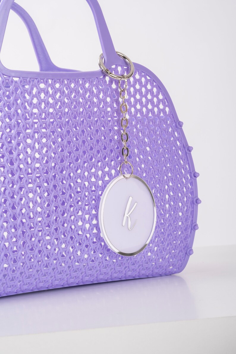 Pu&ograve; includere: Una borsa tote in plastica color lavanda con un motivo a intreccio e una catena argentata con un ciondolo circolare bianco con una lettera "K" argentata.