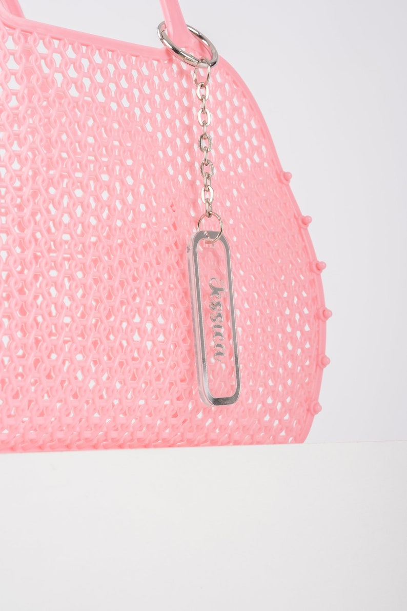 Pu&ograve; includere: Borsa tote in rete rosa con una catena argentata e un portachiavi in acrilico trasparente con la scritta "Jessica" stampata.