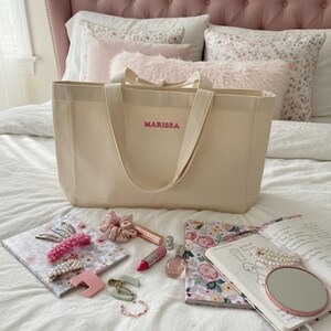 Op de afbeelding: Een beige draagtas met de naam "MARISSA" in roze letters. De tas ligt op een bed met roze kussens en een notitieboek met bloemenpatroon, haaraccessoires, lippenstift, parfum en een kleine spiegel.