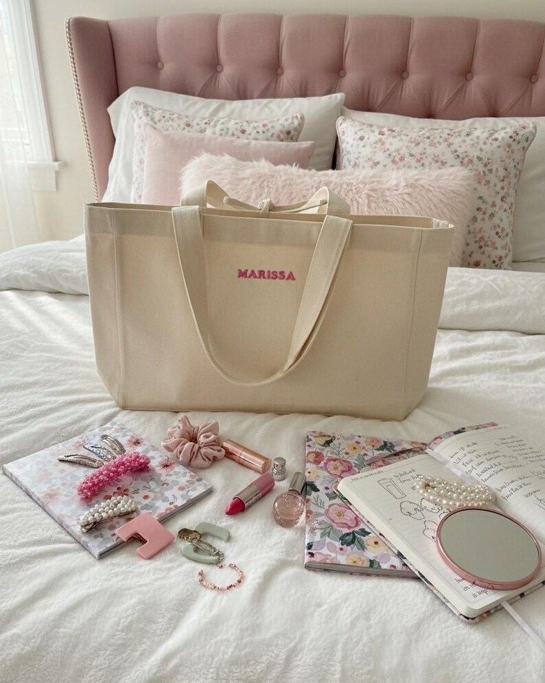 Op de afbeelding: Een beige draagtas met de naam "MARISSA" in roze letters. De tas ligt op een bed met roze kussens en een notitieboek met bloemenpatroon, haaraccessoires, lippenstift, parfum en een kleine spiegel.
