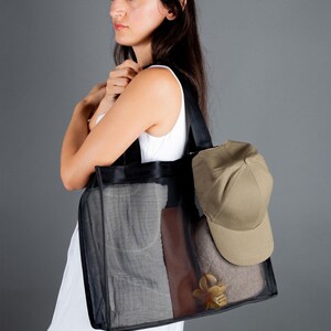 May include: A woman is holding a black mesh tote bag with a brown leather accent. The bag is filled with items, including a beige baseball cap.
