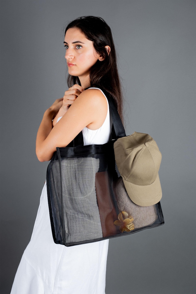 May include: A woman is holding a black mesh tote bag with a brown leather accent. The bag is filled with items, including a beige baseball cap.