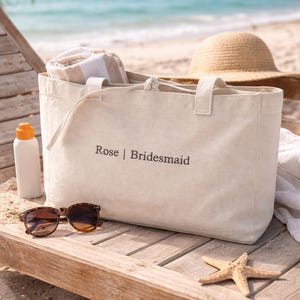 May include: A beige canvas tote bag featuring the text Rose | Bridesmaid printed in dark lettering on the front. The bag has two top handles and a drawstring closure, containing a folded white and beige striped towel inside the main compartment.