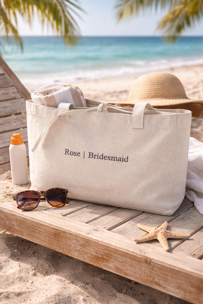 May include: A beige canvas tote bag featuring the text Rose | Bridesmaid printed in dark lettering on the front. The bag has two top handles and a drawstring closure, containing a folded white and beige striped towel inside the main compartment.