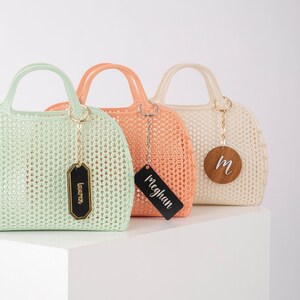 Pu&ograve; includere: Tre borse a rete colorate con etichette in legno personalizzate. Le borse sono verde chiaro, pesca e bianco. Le etichette riportano "Lauren", "Meghan" e "M".